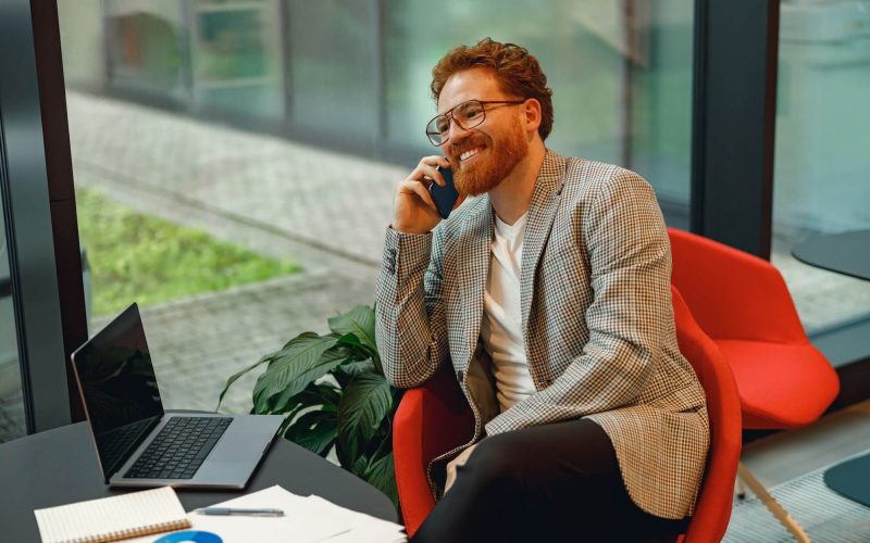 smiling-male-boss-talking-phone-with-client-while-sitting-in-coworking-space-and-working-on.jpg